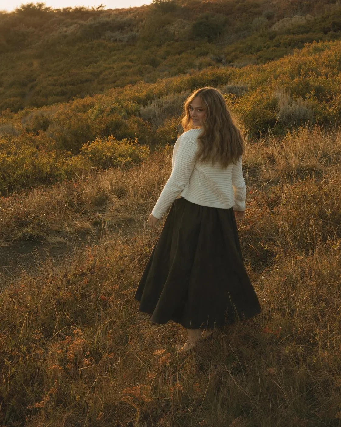 Aliza walking away through a golden autumn field at sunset, glancing back over her shoulder.
