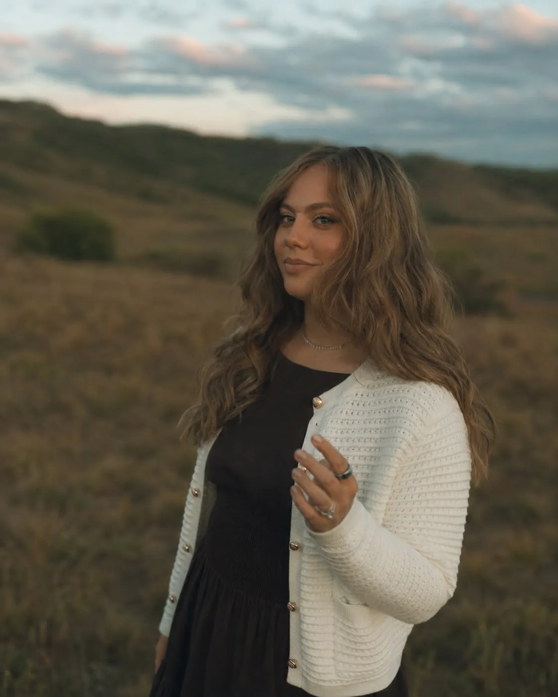 Aliza standing in a golden field at dusk in a cream cardigan, looking gently toward the camera.